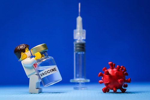 تصویری از یک کاراکتر اسباب بازی لوگو که واکسن کووید 19 در دست دارد و روبرویش یک ویروس کرونا قرار دار