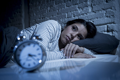 تصویر خانمی در رختخواب که از بی خوابی رنج میبرد و خوابش نمیبرد و یک ساعت در جلو او قرار دارد