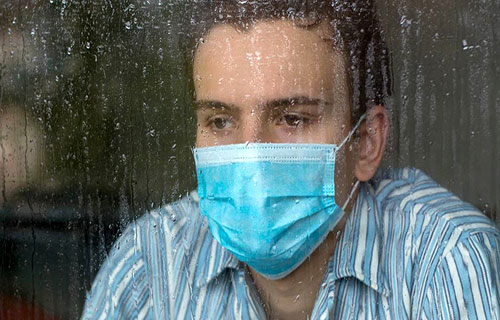 مردی که ماسکپزشکی به صورت دارد و دارد از پنجره به بیرون که باران می آید نگاه میکند
